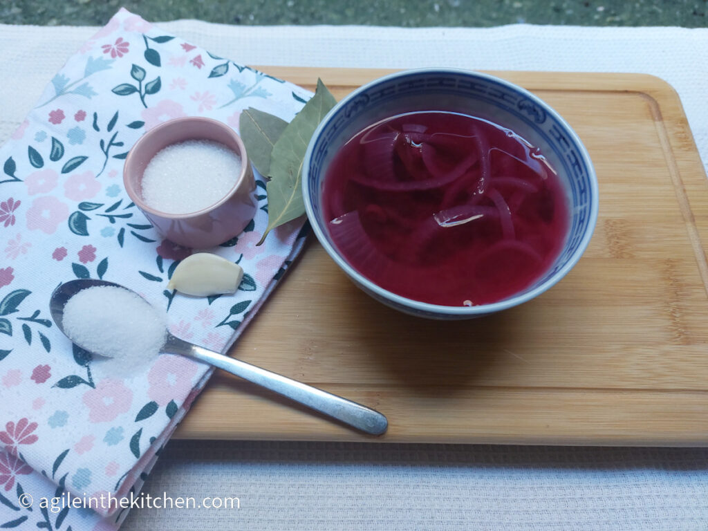 On a wooden cutting board, a flowery cloth napkin folded, on top a teaspoon with salt, a pink jar with sugar, a garlic clove and two bay leaves. Next to the cloth, a ceramic bowl with sliced red onion in pickle juice