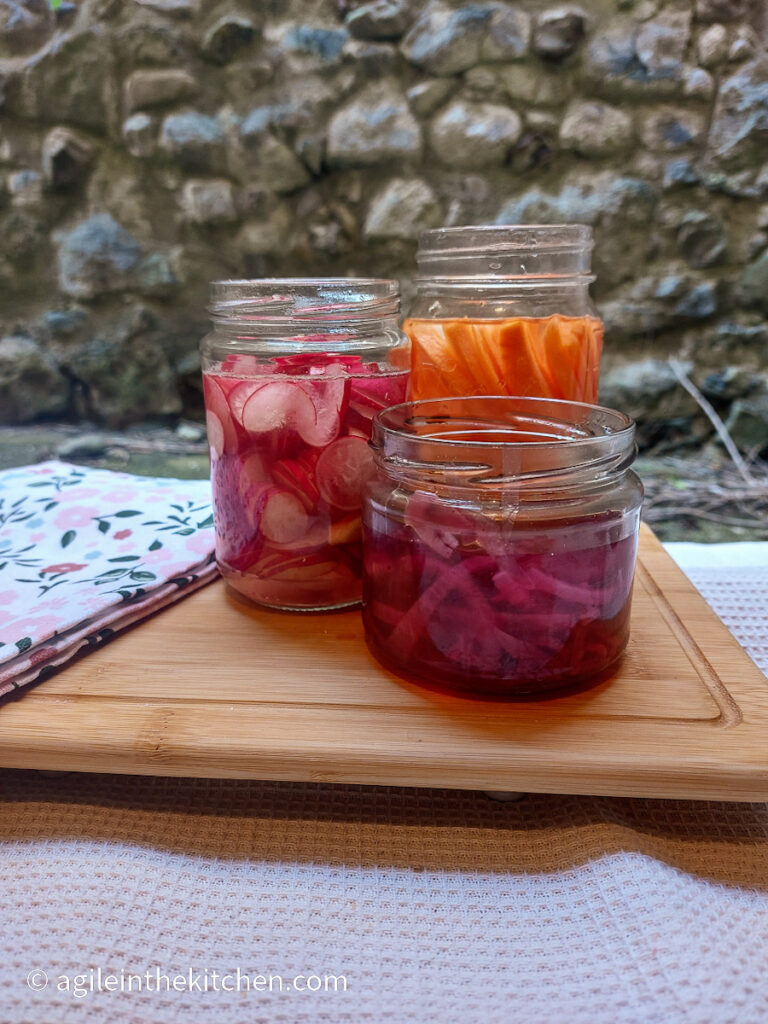 On a wooden cutting board, with a folded flowery cloth napkin, three jars of pickled vegetables; one with sliced radishes, one with carrot sticks and in the front, a jar of sliced red onions.