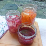 Photo taken slightly form above. A wooden cutting board, with a folded flowery cloth napkin, three jars of pickled vegetables; one with sliced radishes, one with carrot sticks and one with sliced red onions.