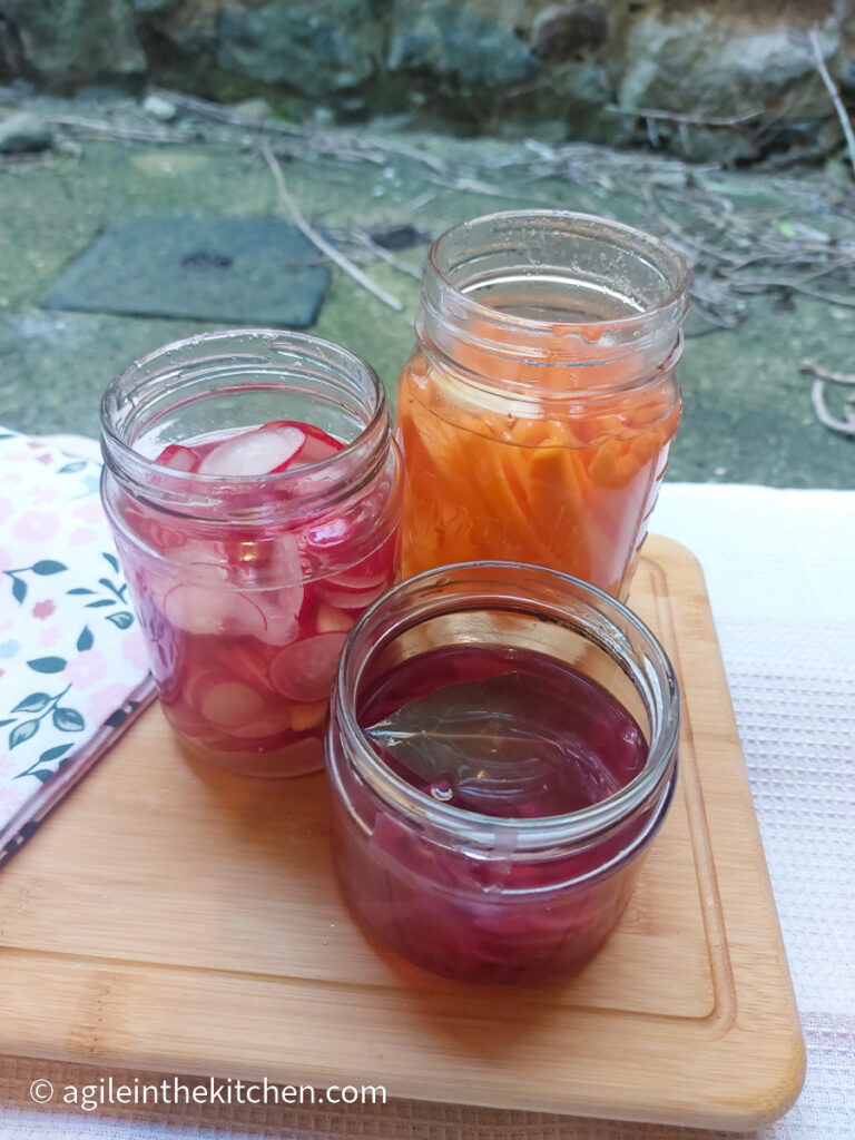 Photo taken slightly form above. A wooden cutting board, with a folded flowery cloth napkin, three jars of pickled vegetables; one with sliced radishes, one with carrot sticks and one with sliced red onions.