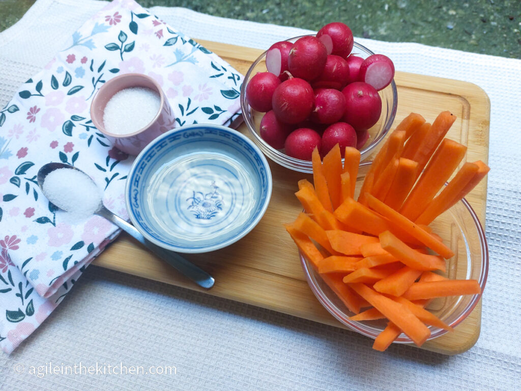 On a wooden cutting board with a folded cloth napkin with flowers, a teaspoon with salt, a pink bowl with sugar, a blue ceramic bowl with vinegar, a glass bowl with radishes and a glass bowl with carrot sticks