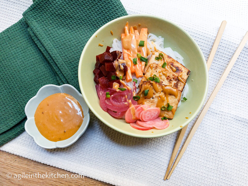 On a white textured background, a green folded cloth napkin in the left corner, towards the right corner a ceramic bowl shaped as a flower filled with Banh mi sauce, in the middle of the picture a green bowl with rice noodles, pickled vegetables, baked tofu and sprinkle of green onions, to the right a set of wooden chop sticks