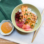 On a white textured background, a green folded cloth napkin in the left corner, towards the right corner a ceramic bowl shaped as a flower filled with Bahn mi sauce, in the middle of the picture a green bowl with rice noodles, pickled vegetables, baked tofu and sprinkle of green onions, to the right a set of wooden chop sticks