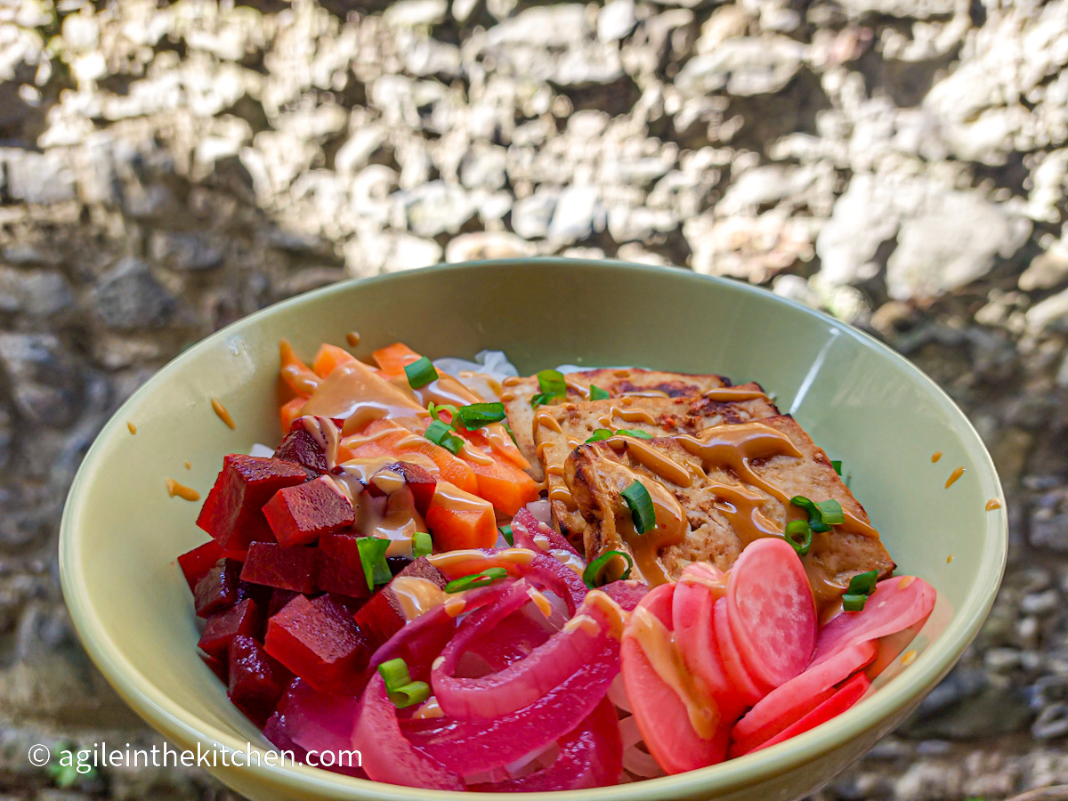 A green bowl with tofu bahn mi; rice noodles, pickled vegetables, baked tofu, spring onions, and spicy sauce.