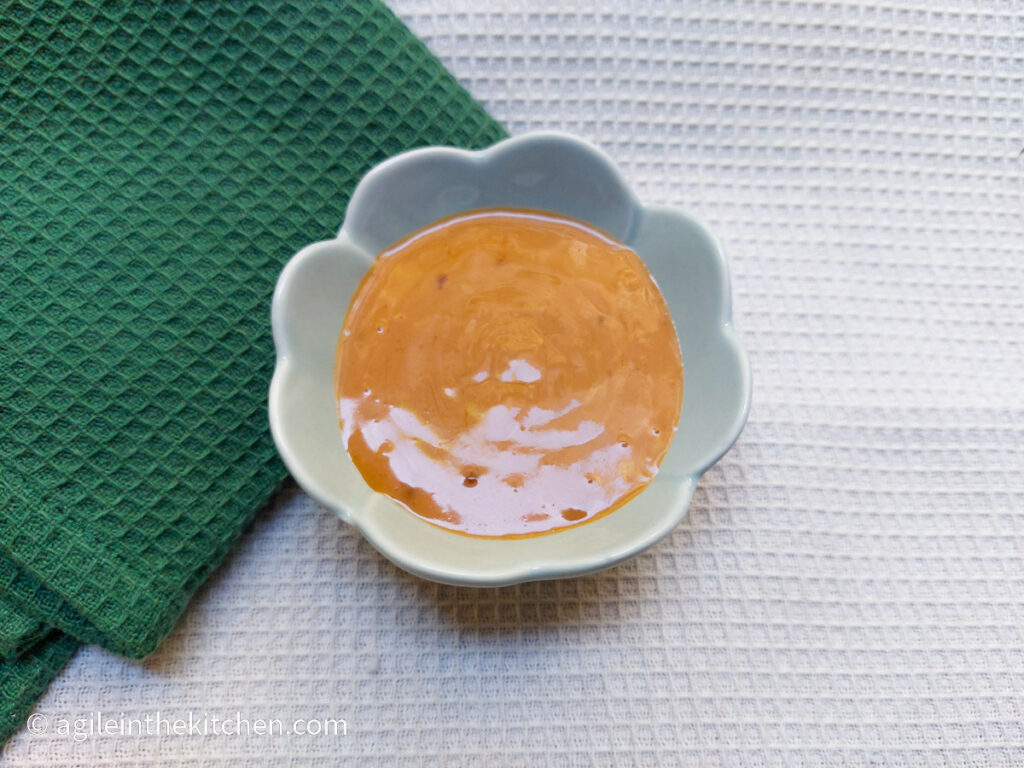 On a white textured background, in the middle of the pictures a blue, porcelain bowl shaped like a flower filled with bahn mi sauce, to the left a textured green cloth napkin folded.