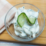 On a wooden cutting board background with a pink plaid cloth napkin in the left corner, a glass bowl with tzatziki with a sliced cucumber twisted on top.