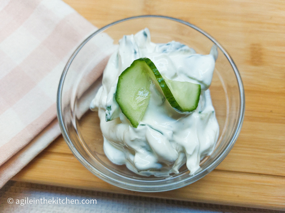 On a wooden cutting board background with a pink plaid cloth napkin in the left corner, a glass bowl with tzatziki with a sliced cucumber twisted on top.