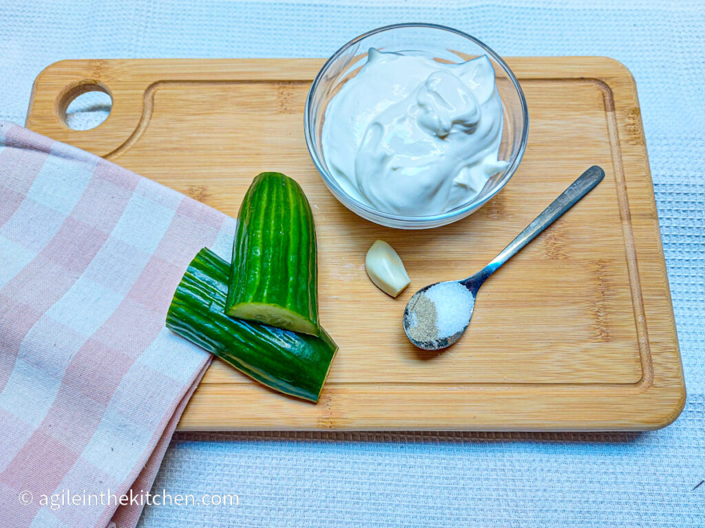 On a wooden chopping board, a folded pink plaid cloth napkin, a piece of cucumber cut lengthwise, a glass bowl of Greek yogurt, a clove of garlic and a teaspoon with salt and white pepper