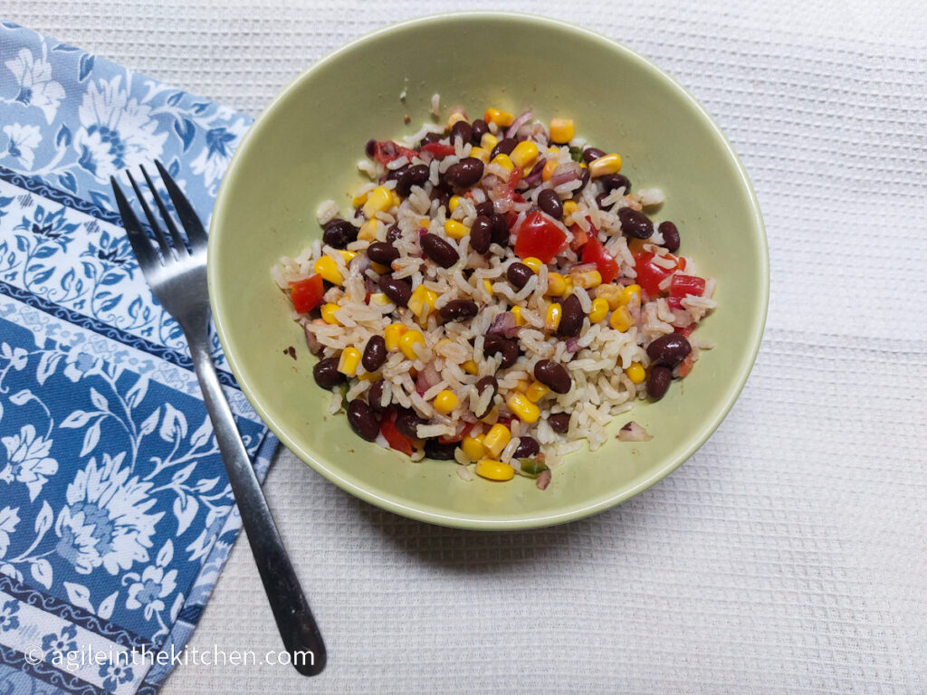 On a white textured background, a green bowl in the middle of the photo with black bean salsa mixed with rice, next to the plate is a fork and folded underneath the fork and plate is a blue cloth napkin with a white flower pattern.