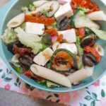 A blue bowl with Mediterranean pasta salad, sitting on top of a cloth with a flower pattern.