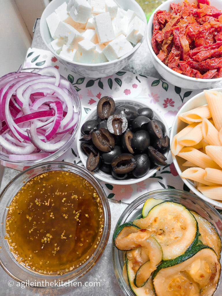 In bowls ingredients for a Mediterranean pasta salad. Clockwise from top; feta cheese diced, sun dried tomatoes sliced, penne pasta, zucchini sliced, vinaigrette dressing, red onion sliced, and in the middle black olives halved.