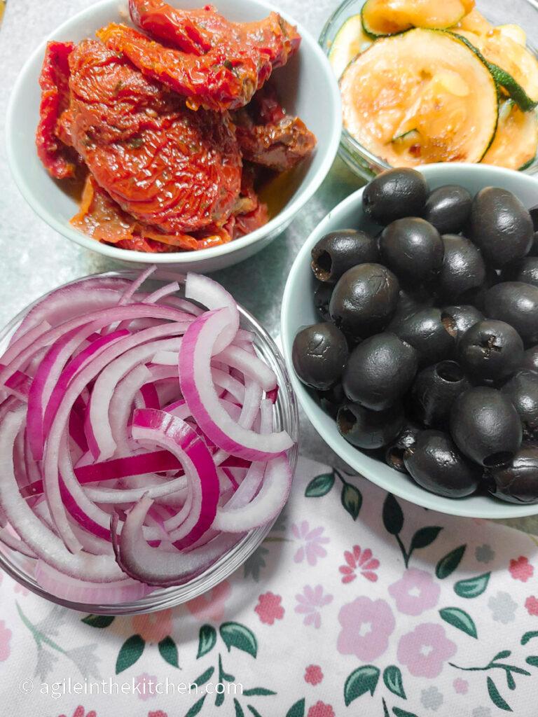 In bowls ingredients for a Mediterranean pasta salad. Clockwise from top left; sun dried tomatoes, zucchini sliced, black olives and red onions sliced.