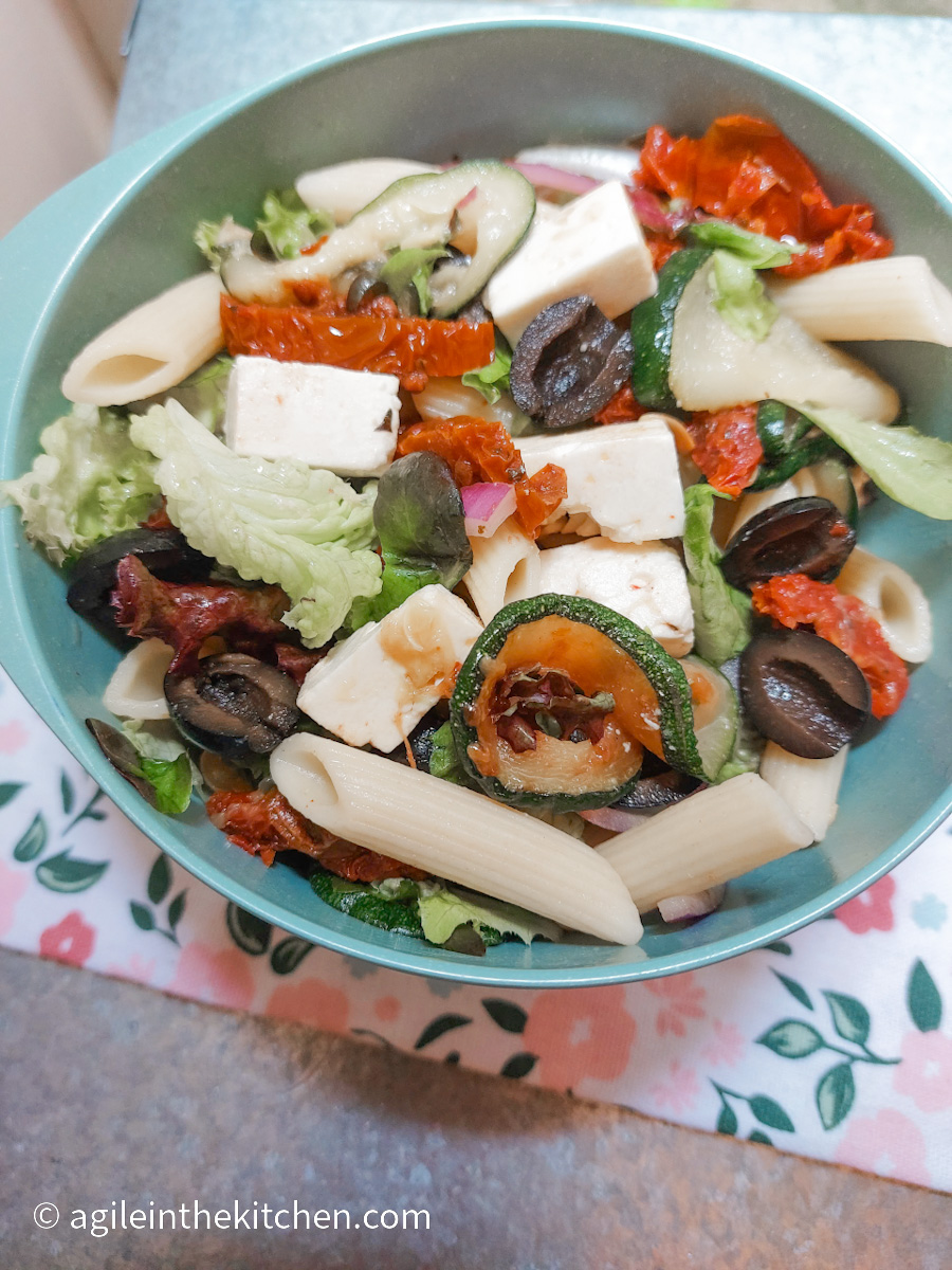 A blue bowl with Mediterranean pasta salad, sitting on top of a cloth with a flower pattern.