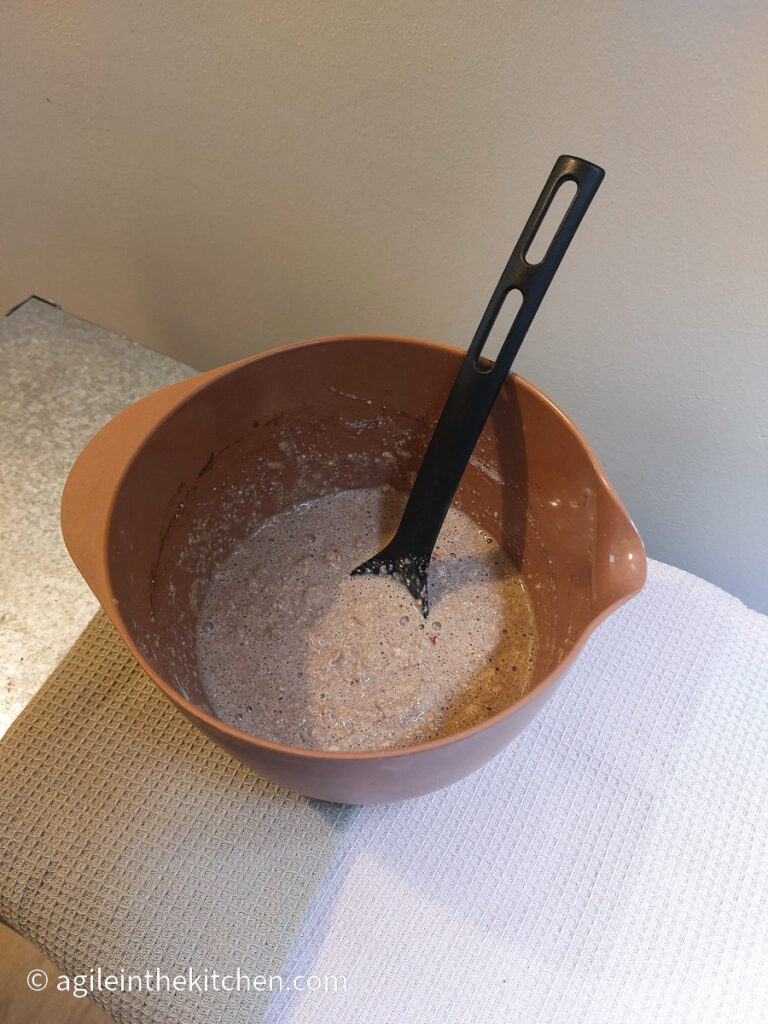 On a textured background, an orange bowl with a spoon and pancake batter