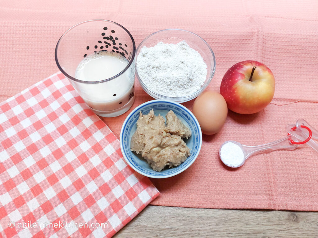 On a pink textured background, a red and white gingham cloth in the lower left corner, in the middle a glass of milk, a bowl of flour, an egg, an apple, a teaspoon filled with baking powder and a small bowl of peanutbutter.