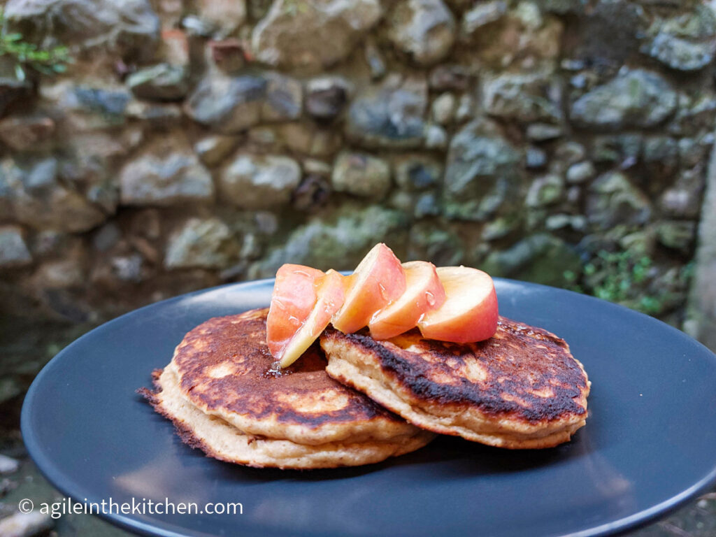 On a textured background a black matte plate, two pancakes and five slices of apple drizzled with honey.