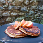 On a textured background a black matte plate, two pancakes and five slices of apple drizzled with honey.