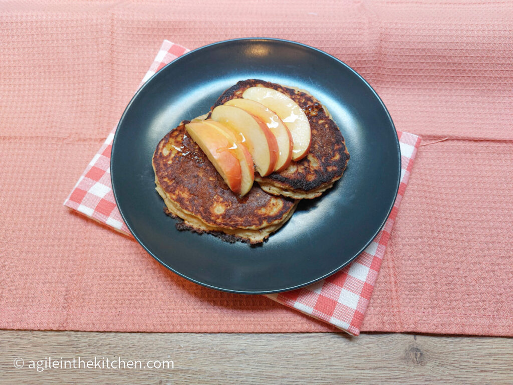 On a deep pink textured background, a black matte plate with a red and white gingham cloth underneath. On the plate two pancakes and five slices of apple drizzled with honey.