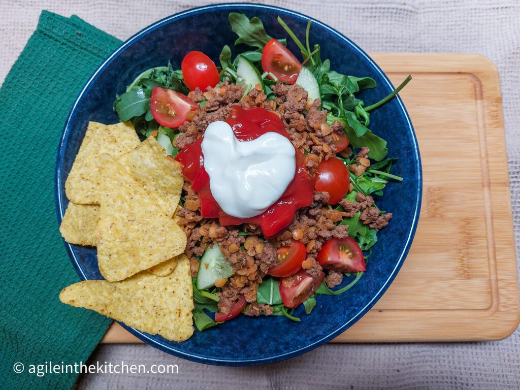 On a wooden cutting board, a blue bowl with taco salad (lettuce, cucumber, tomatoes, taco mince meat with red lentils, salsa and sour cream), with some nacho chips on the side. On the right a folded green cloth napkin