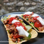 On a black matte plate, two taco tubs filled with taco mince meat, cucumbers, lettuce, salsa and sour cream.