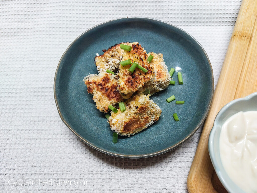 On a white textured background, on a blue plate, aubergine bites in a pile, sprinkled with cut up spring onions. In the right corner, a bowl barely visible with a dipping sauce