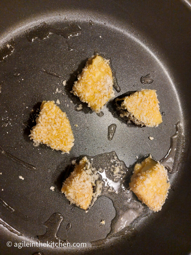 In a black frying pan, five breaded aubergine bites are frying in oil