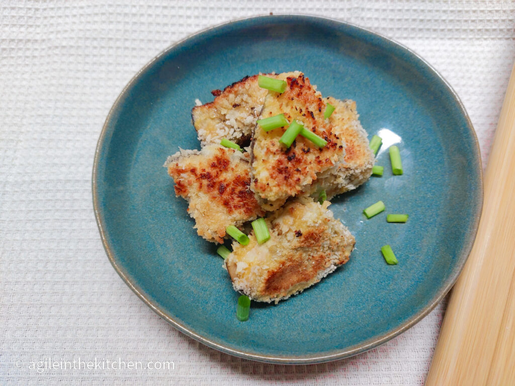 On a white textured background, on a blue plate, aubergine bites in a pile, sprinkled with cut up spring onions.