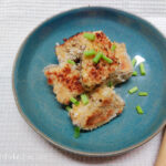 On a white textured background, on a blue plate, aubergine bites in a pile, sprinkled with cut up spring onions.