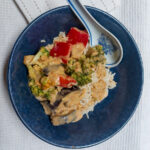 On a white textured background, a blue bowl on a bed of rice, vegetable red curry