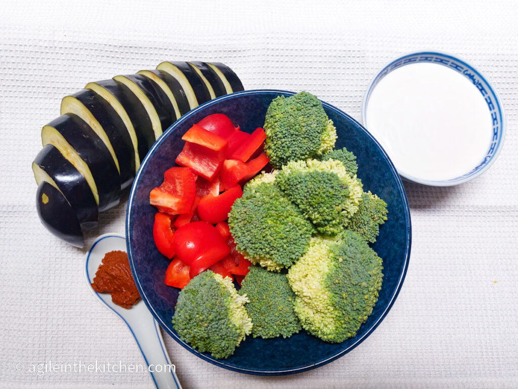 On a white textured background, ingredients for a vegetable curry; one aubergine sliced, in a blue bowl a head of broccoli cut up, one red pepper cut up, a spoonful of red curry paste, a bowl of coconut milk