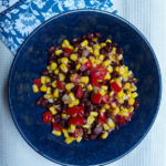 On a white textured background, a blue plate in the middle of the photo with black bean salsa, folded underneath the plate is a blue cloth napkin with a white flower pattern.