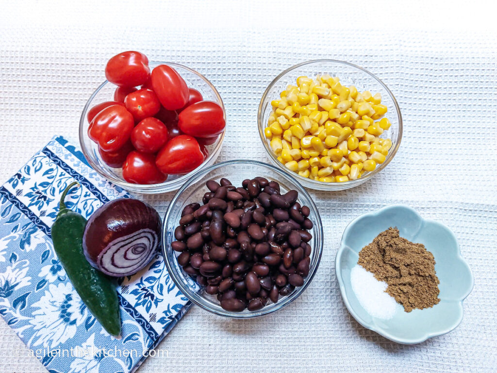 On a white textured background, the ingredients for black bean salsa, with a folded blue cloth napkin in the left hand corner. Clockwise from left; a bowl of plum tomatoes, a bowl of sweet corn, a bowl of salt and cumin, a bowl of black beans, a red onion and a green chili.