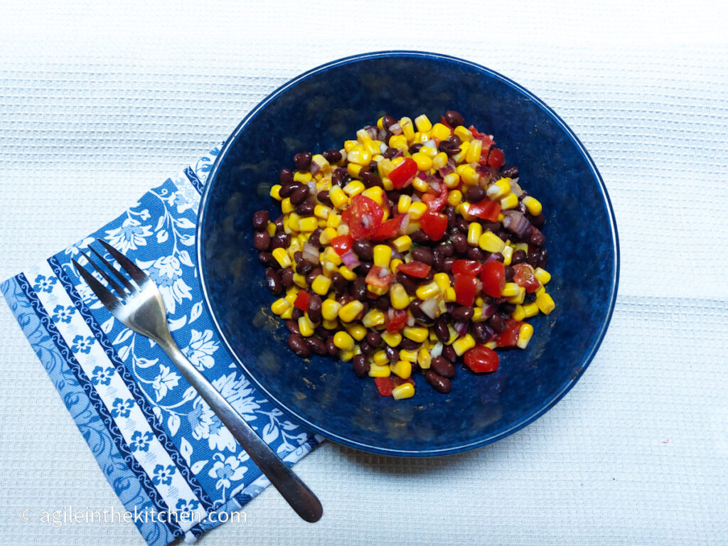 On a white textured background, a blue plate in the middle of the photo with black bean salsa, next to the plate is a fork and folded underneath the fork and plate is a blue cloth napkin with a white flower pattern.