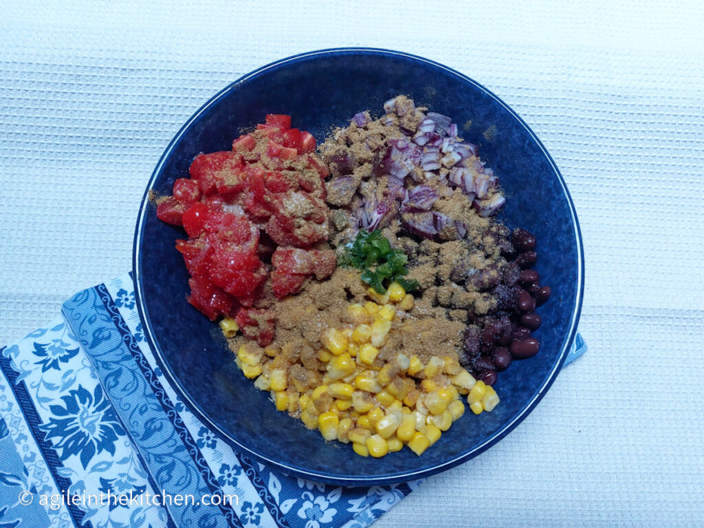 On a white textured background, a blue plate in the middle of the photo with the ingredients for black bean salsa unmixed. Folded underneath the plate is a blue cloth napkin with a white flower pattern.