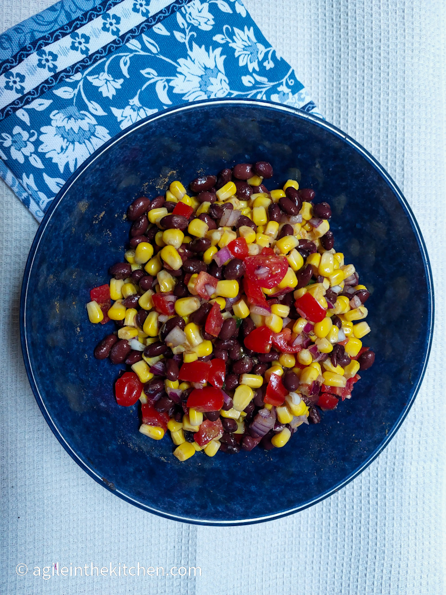 On a white textured background, a blue plate in the middle of the photo with black bean salsa, folded underneath the plate is a blue cloth napkin with a white flower pattern.