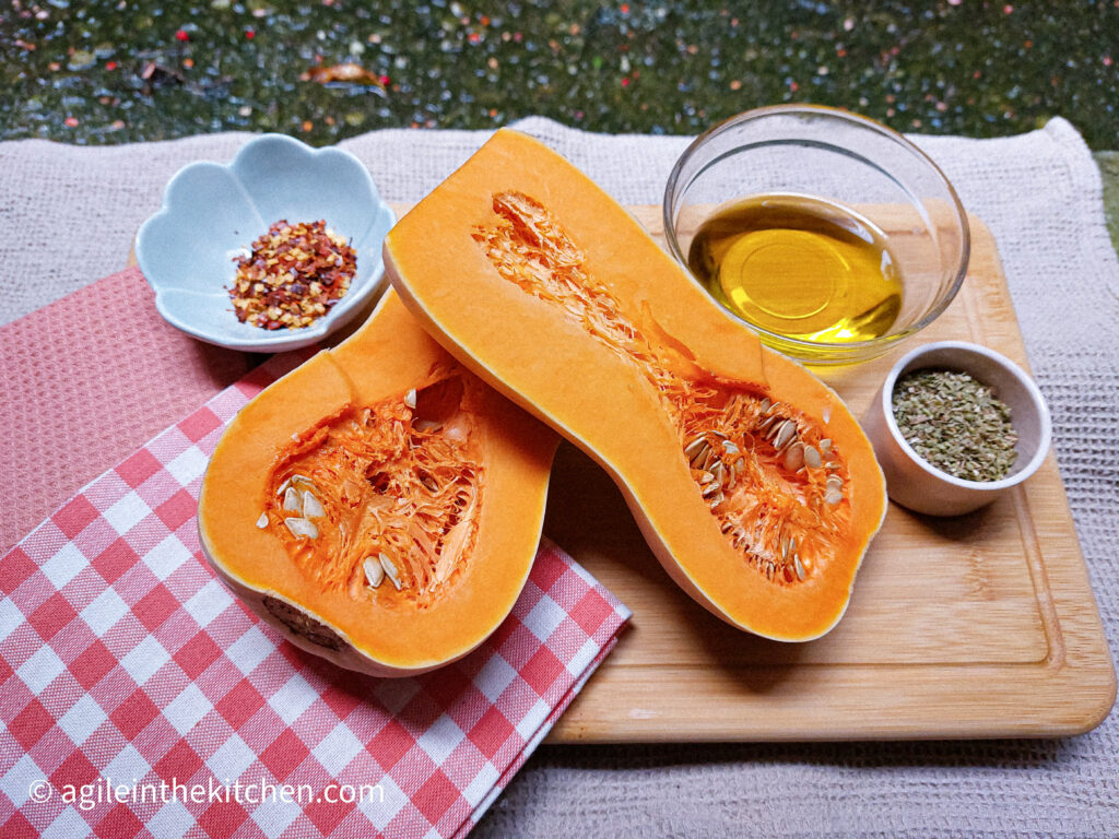 On a wooden cutting board, with a red and white gingham cloth in the right corner a bowl of oil, a butternut squash cut in half, a flower shaped bowl with chili flakes and a bowl of oregano and salt mixed.