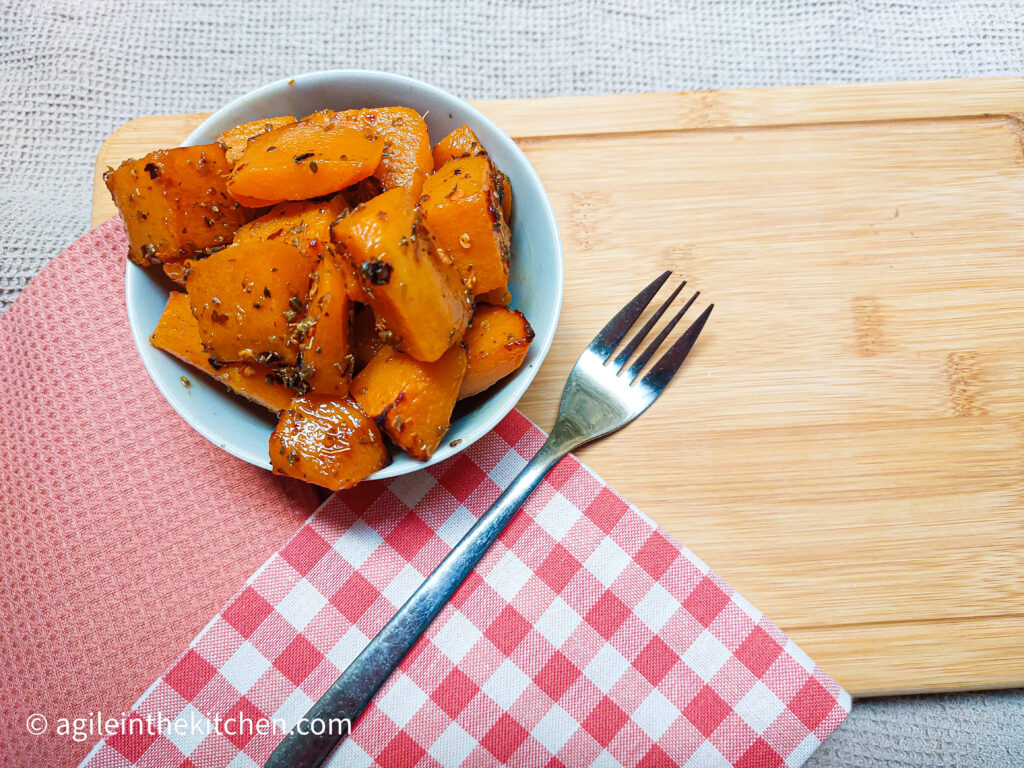 On a wooden cutting board with a red and white gingham cloth and a textured orange cloth folded in the right corner, a bowl of roasted butternut squash next to it, a fork.