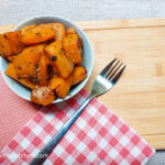 On a wooden cutting board with a red and white gingham cloth and a textured orange cloth folded in the right corner, a bowl of roasted butternut squash next to it, a fork.