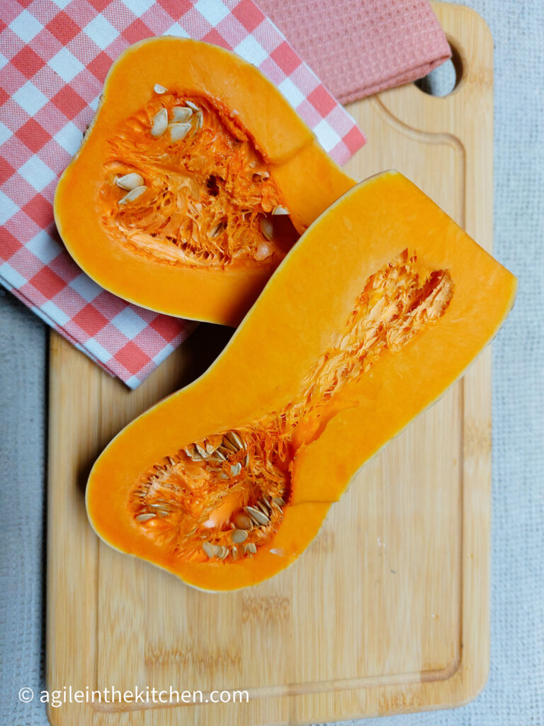 On a wooden chopping board, a folded red and white gingham cloth, a butternut squash cut in half.