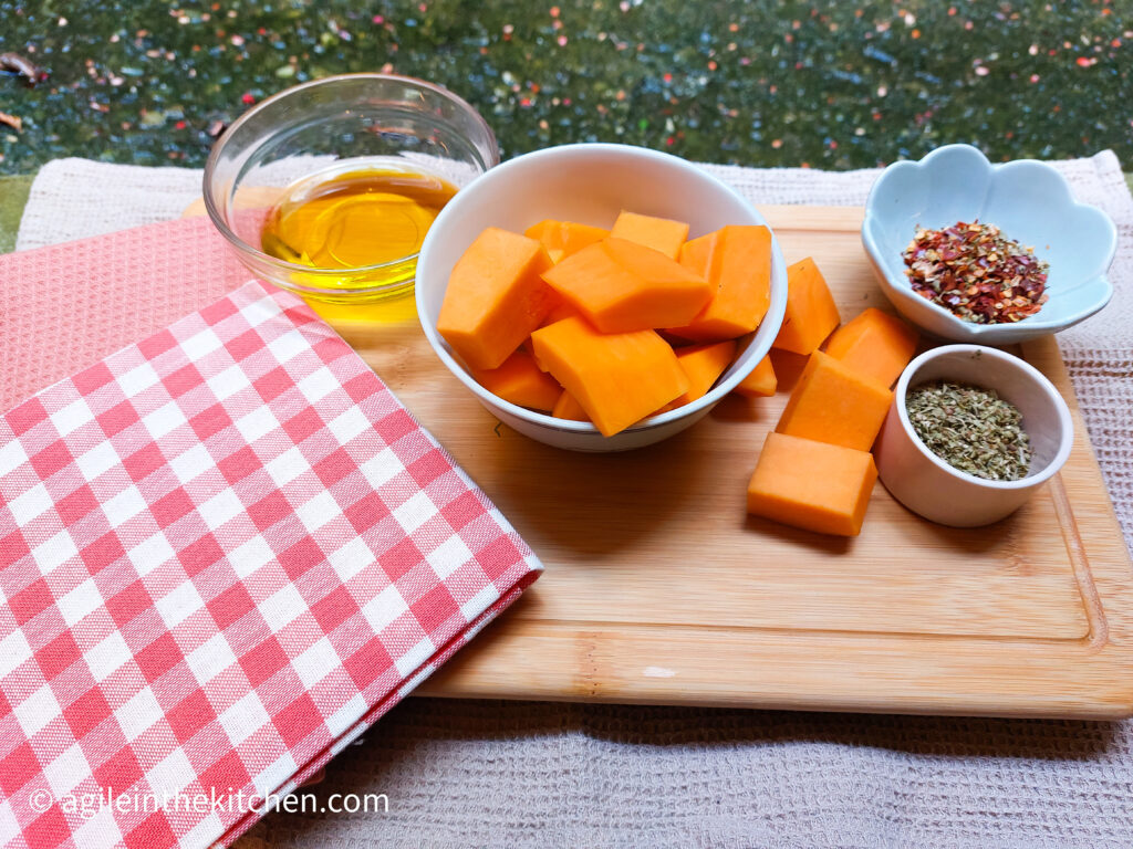 On a wooden cutting board, with a red and white gingham cloth in the right corner a bowl of oil, a bowl of chopped up butter nut squash with some falling over the side, a flower shaped bowl with chili flakes and a bowl of oregano and salt mixed.