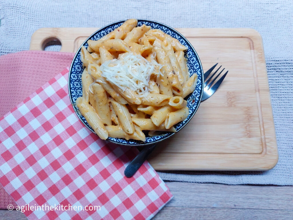 On a wooden cutting board, two folded cloth napkins, one light red and one red gingham. A bowl of butternut squash pure and penne pasta with some shredded cheese on top.