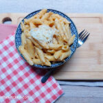 On a wooden cutting board, two folded cloth napkins, one light red and one red gingham. A bowl of butternut squash pure and penne pasta with some shredded cheese on top.