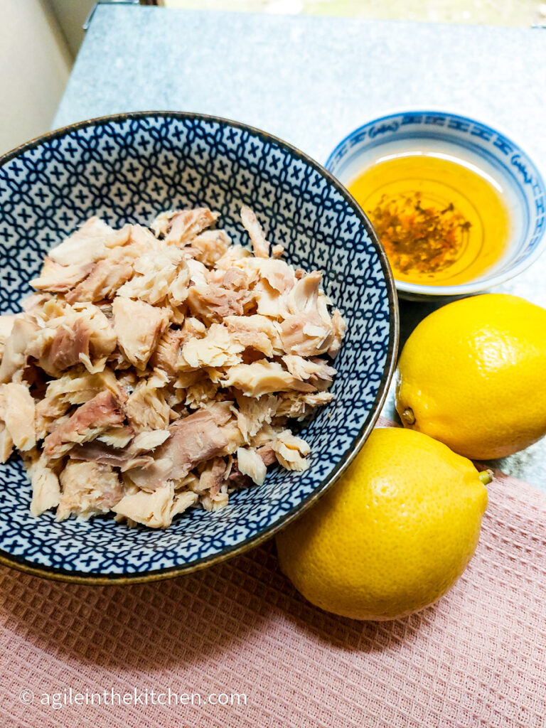 A blue patterened bowl with flaked salmon, a small bowl with olive oil and two lemons.