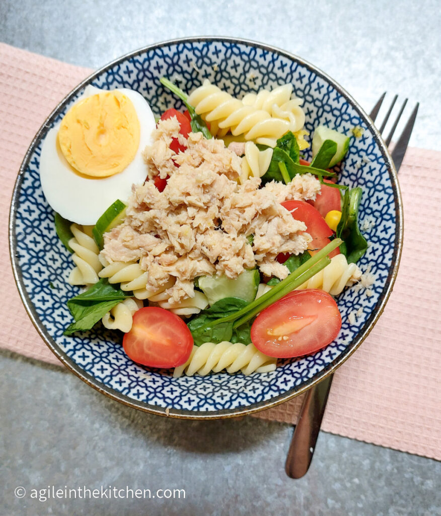 A blue patterned bowl with lemon salmon pasta salad, underneath the bowl a pink, textured cloth and a fork