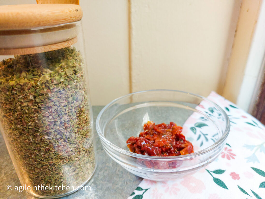 A glass jar filled with oregano, next to a glass bowl with sin dried tomatoes cut up