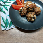 A white cloth with green leaves folded underneath a black matte plate with a pile of Mediterranean meatballs and a tomato cut in wedges.