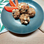 A white cloth with green leaves folded underneath a black matte plate with a pile of Mediterranean meatballs and a tomato cut in wedges.