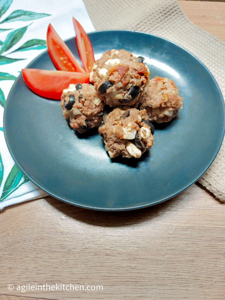 A white cloth with green leaves folded underneath a black matte plate with a pile of Mediterranean meatballs and a tomato cut in wedges.