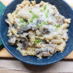 On a wooden cutting board, a green cloth folded, a blue pasta bowl with creamy mushroom pasta.