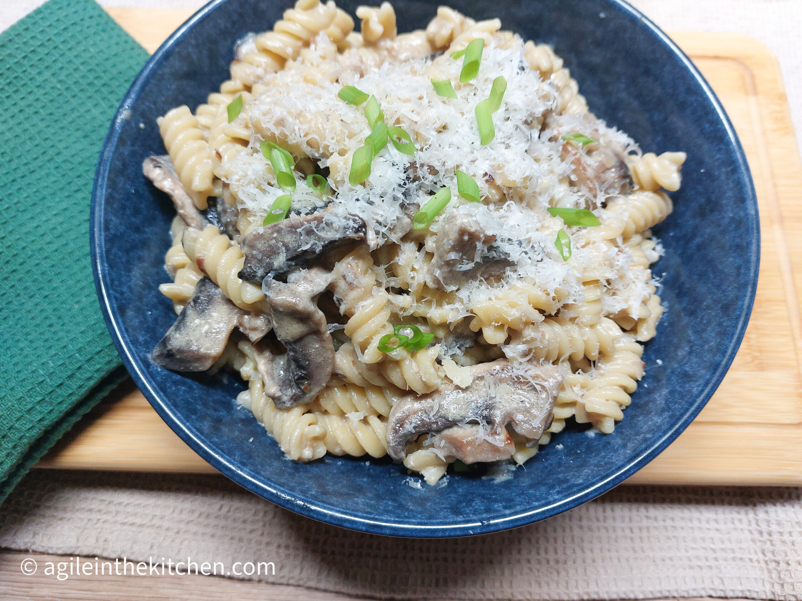 On a wooden cutting board, a green cloth folded, a blue pasta bowl with creamy mushroom pasta.